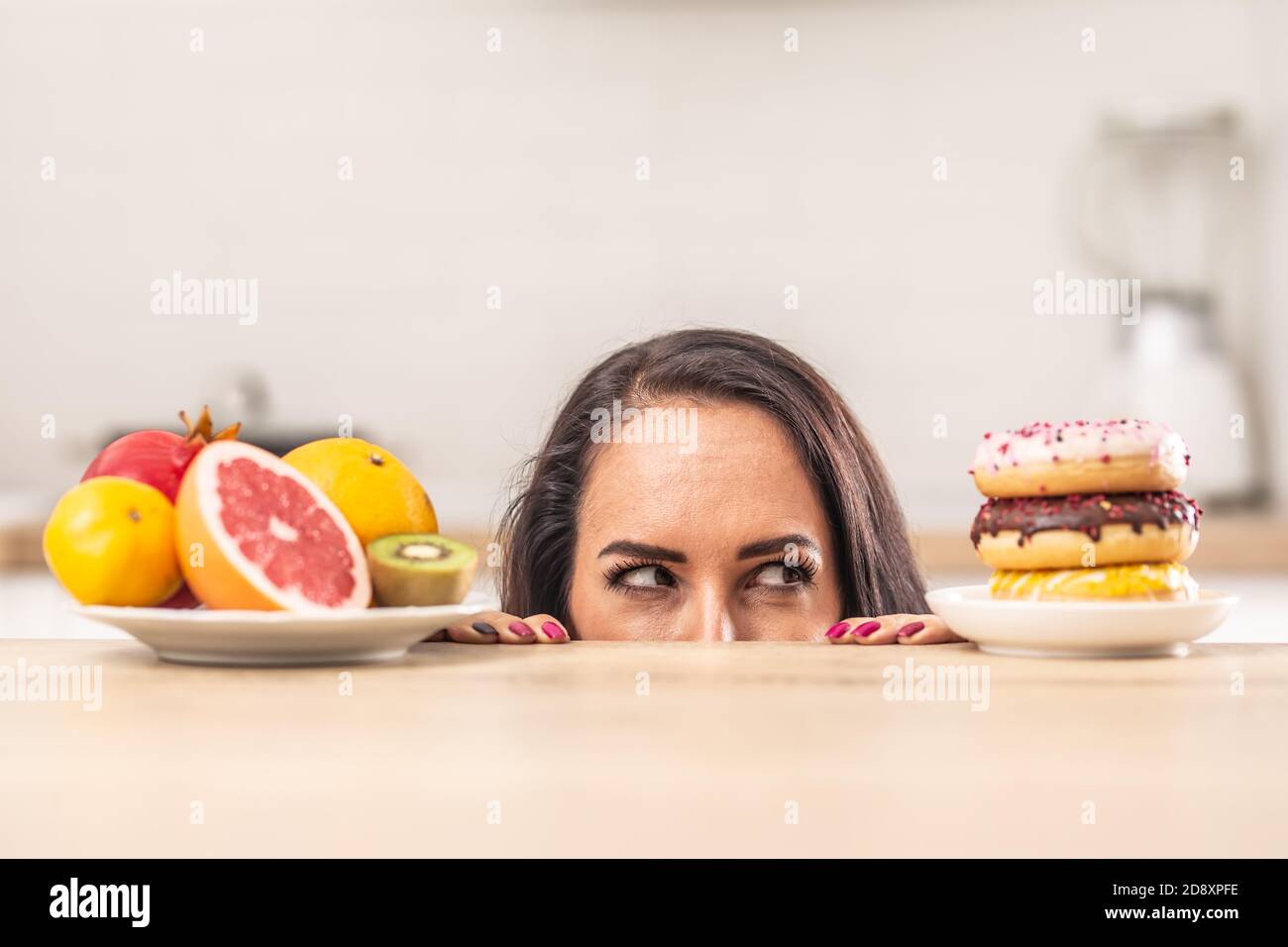 Weibliche Blicke auf Donuts stapeln sich auf einem Tisch als gegenüber einem Teller voller frischer Früchte. Stockfoto