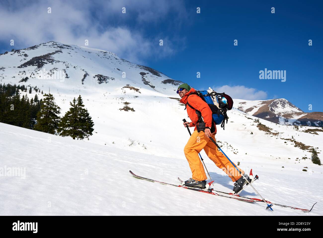 Skifahrer Tourist Backpacker, glücklich lächelnd bärtigen Mann Wandern auf Skiern Hügel auf dem Hintergrund des blauen Himmels und schöne Bergkämme. Winterurlaub, aktiver Lifestyle, Ski- und Trekkingkonzept. Stockfoto