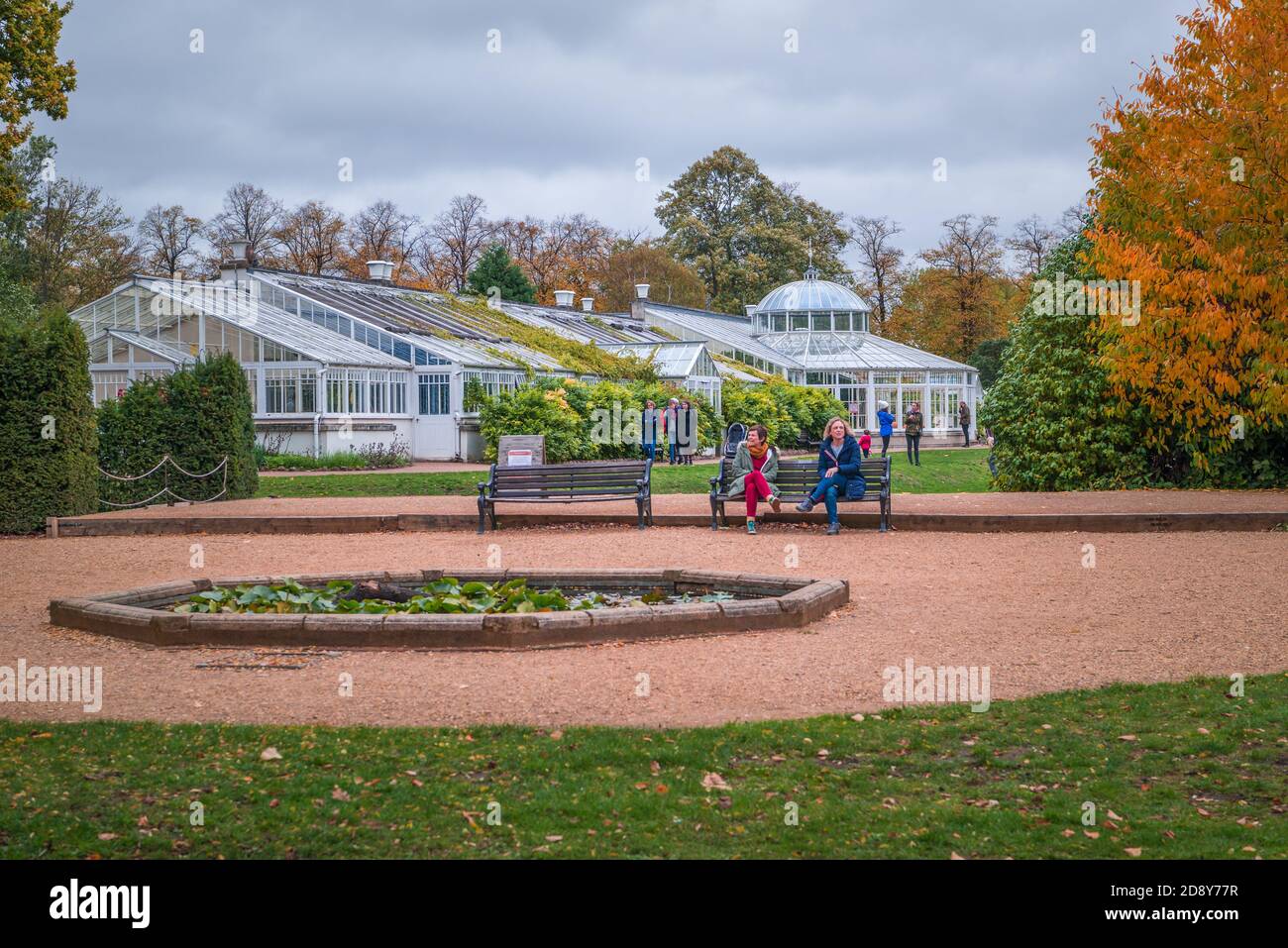 Menschen im Chiswick House Garten im Herbst. Zwei Frauen sitzen auf einer Bank mit Gewächshäusern im Hintergrund. London, England, Großbritannien Stockfoto