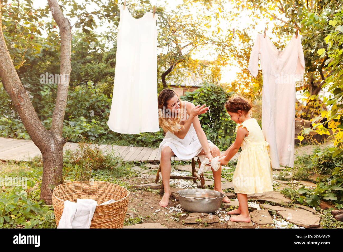 Charming kleines Mädchen helfen Mutter mit Weidenkorb während der Arbeit Chore und hängende Wäsche im Hinterhof im Sommer Abend Stockfoto