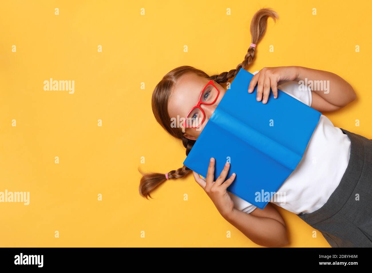 Draufsicht auf eine kleine Schülerin mit Brille auf gelbem Hintergrund. Das Kind versteckt sich hinter einem Buch. Stockfoto