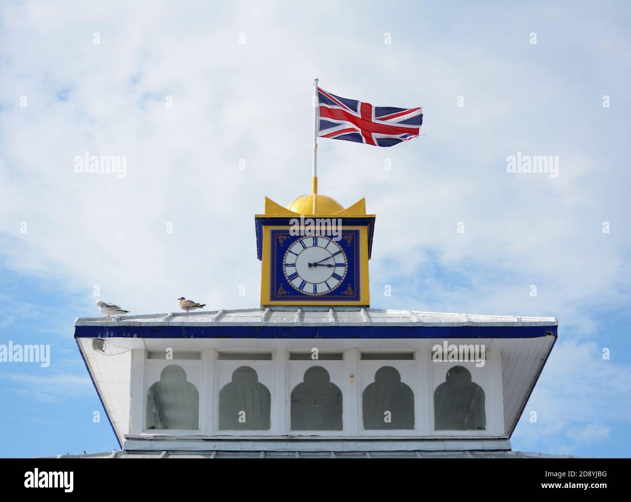 Die Union Jack Flagge fliegt auf dem Uhrenturm des Eastbourne Pier. Möwen sitzen bei Sonnenschein auf dem Dach. Stockfoto