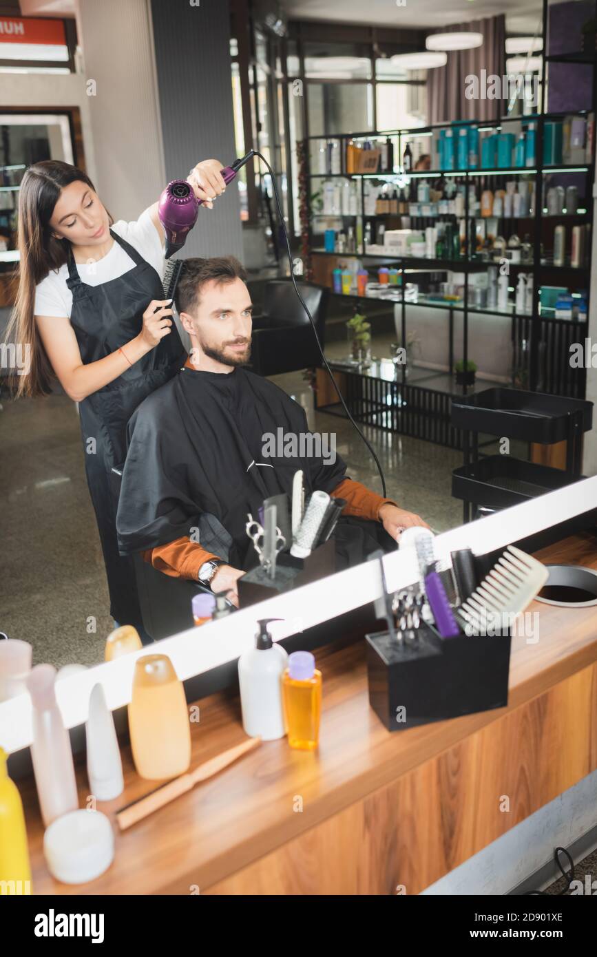 Spiegelreflexion der Friseur Trocknen Haar des Kunden mit kosmetischen Flaschen auf verschwommenem Vordergrund Stockfoto