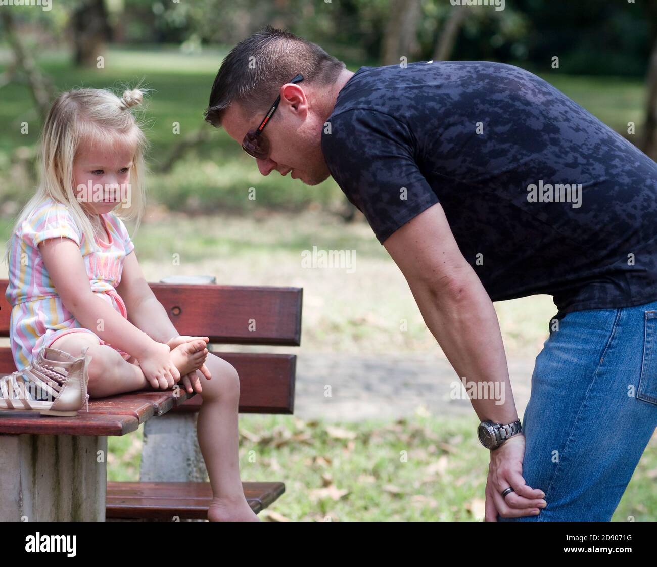 Ein Vater zeigt sich besorgt um seine traurige Tochter in einem Park und hockt auf ihre Ebene, während sie auf einem Tisch sitzt und ihren Fuß hält. Stockfoto