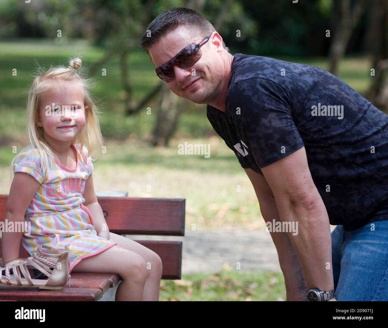 Auf einem Ausflug in den Park sitzt eine Tochter auf einem Tisch und lächelt an der Kamera, während ihr Vater auf ihre Ebene hockt Stockfoto