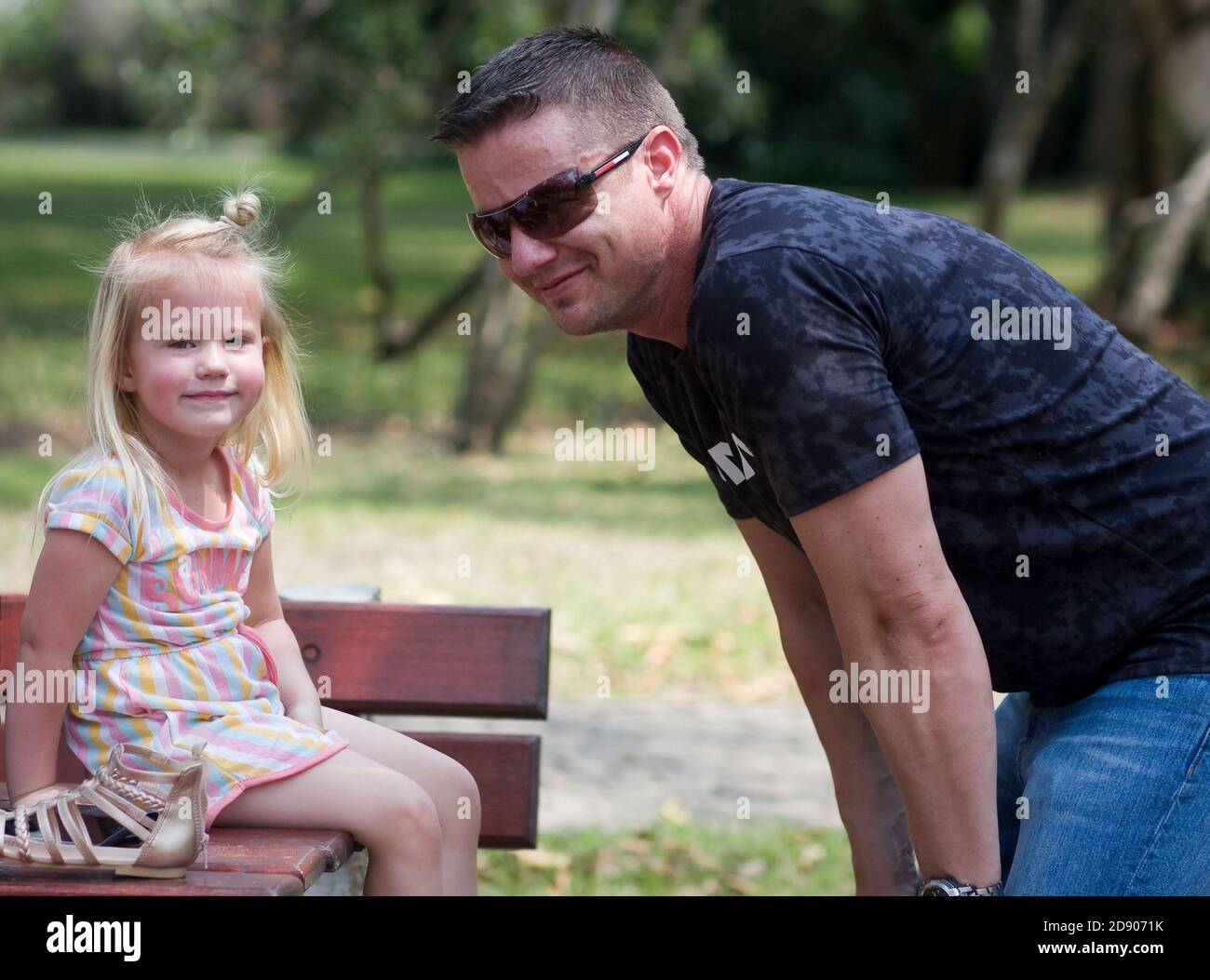 Auf einem Ausflug in den Park lächelt eine Tochter die Kamera an, während ihr Vater sie stolz anlächelt und sich auf ihr Niveau hockend. Stockfoto
