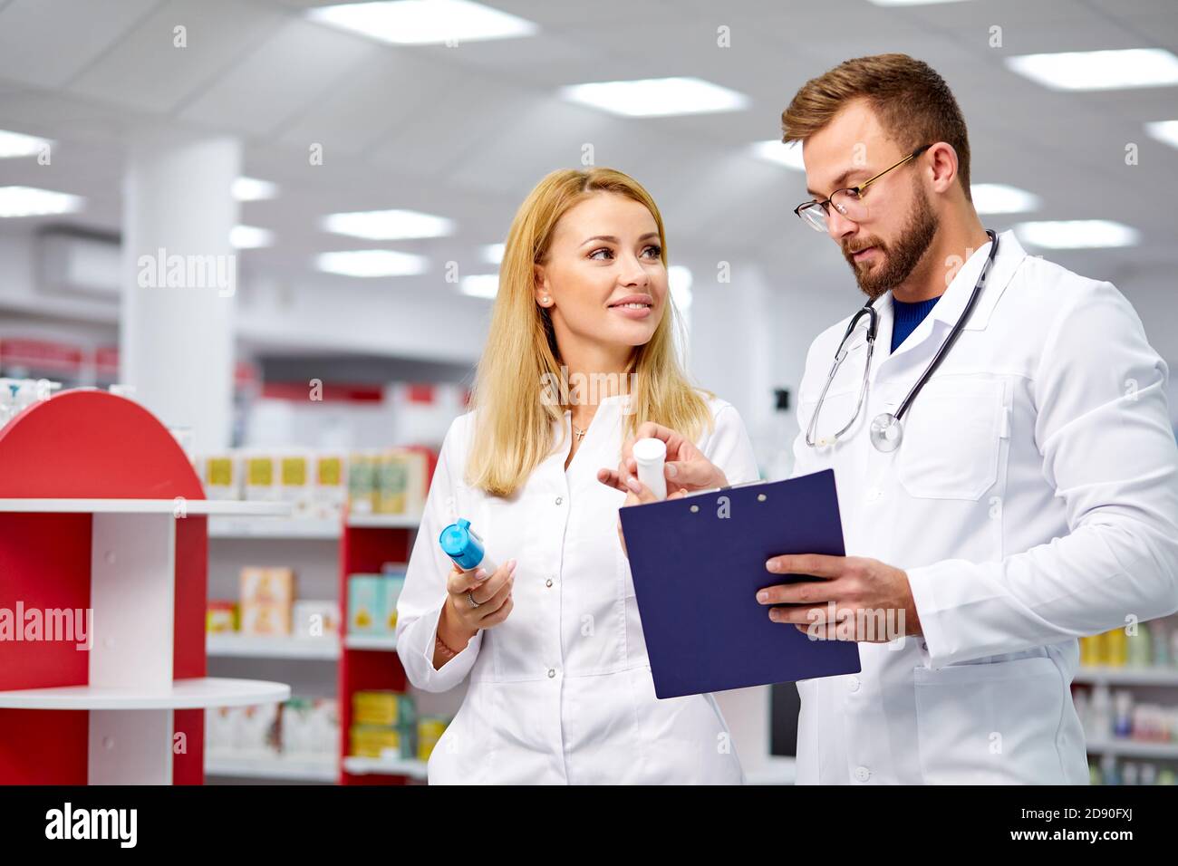 Zwei junge kaukasische Apotheker diskutieren Medikamente, mit einer digitalen Tablette und Dokumente. Mann und Frau in weißem medizinischen Kleid arbeiten in der Apotheke Stockfoto
