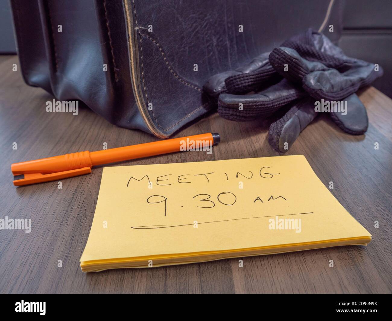 Nahaufnahme ‘meiner alten Ledertasche und einem Paar Handschuhe auf einem Schreibtisch, mit einem Stift und einer Nachricht, die „9.30 Uhr früh“ bestätigt. Stockfoto