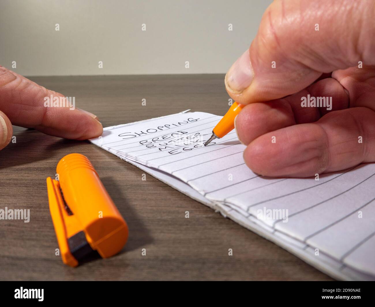 Nahaufnahme eines Mannes mit der Hand, der einen orangefarbenen Filzstift hält und eine Einkaufsliste auf einem linierten Notizblock auf einem Schreibtisch schreibt. Stockfoto
