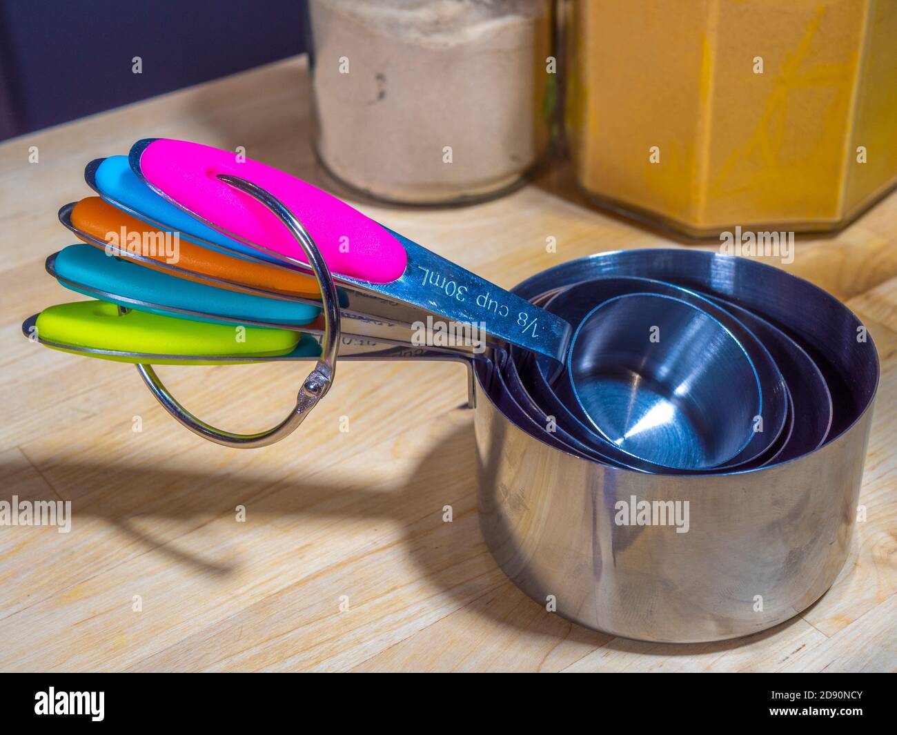 Ein Satz von Messbecher aus Edelstahl unterschiedlicher Größe, mit farbcodierten Griffen, auf einer Arbeitsfläche in der Küche, mit Glasgefäßen mit Zutaten dahinter. Stockfoto