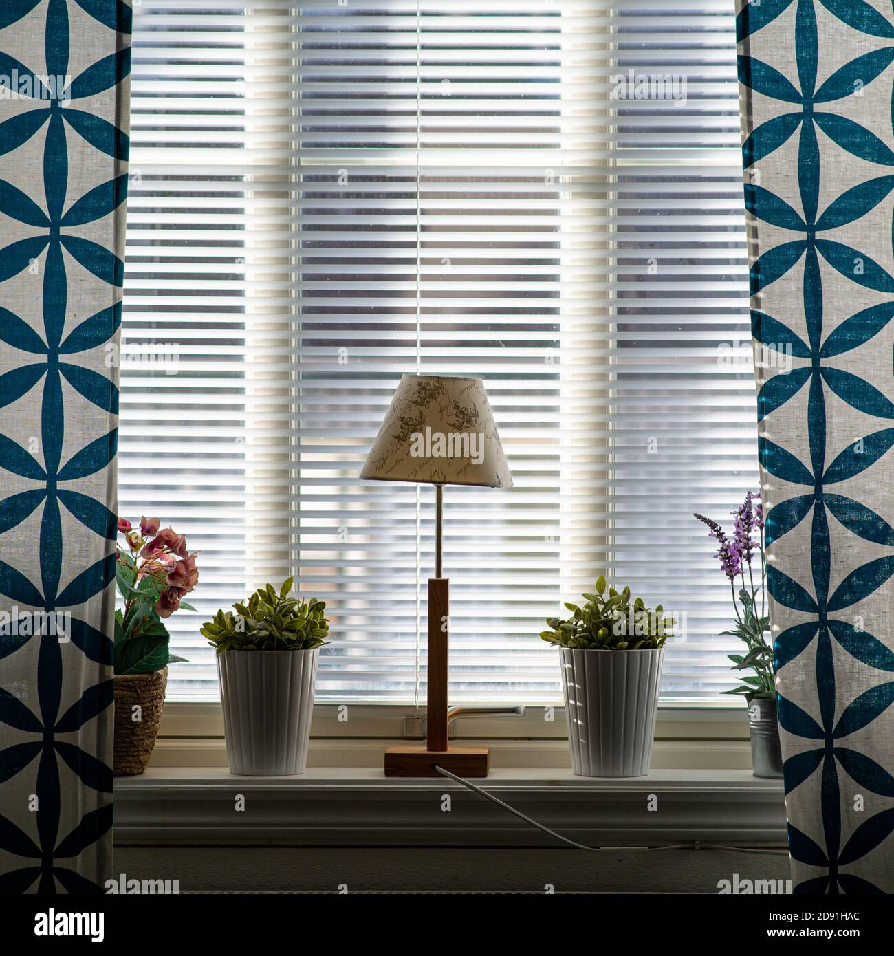 Ein Fenster mit blauen Vorhängen, einer Lampe und Blumen. Schönes Licht kommt durch die Jalousien Stockfoto