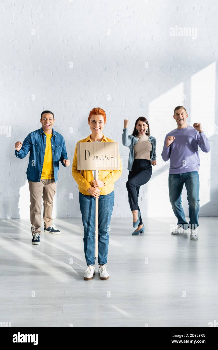 Glückliche Frau hält Plakat mit Demokratie Schriftzug in der Nähe glücklich multikulturell Menschen tanzen im Hintergrund Stockfoto