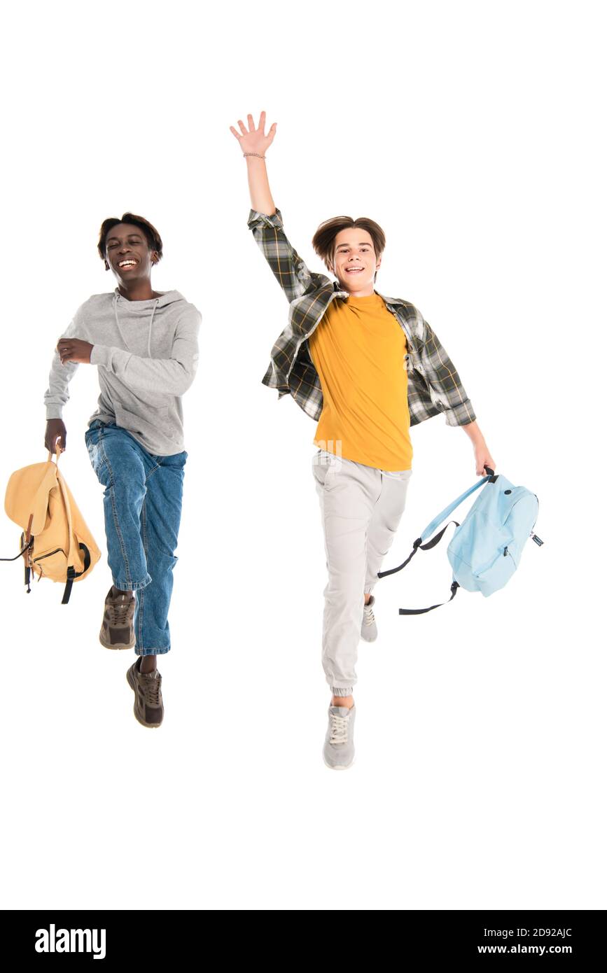 Fröhliche multiethnische Teenager mit Rucksäcken springen isoliert auf weiß Stockfoto