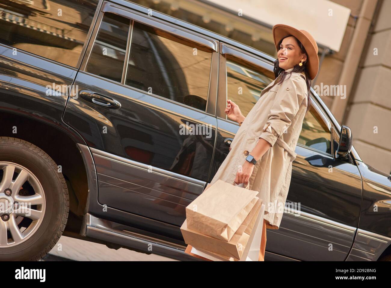 Abschluss des Einkaufs. Junge glücklich stilvolle Frau mit Papier Einkaufstaschen, Blick beiseite und lächelnd, während sie gegen schwarzen Luxus-Auto auf der Straße der Stadt. Menschen Lifestyle, Mode Stockfoto