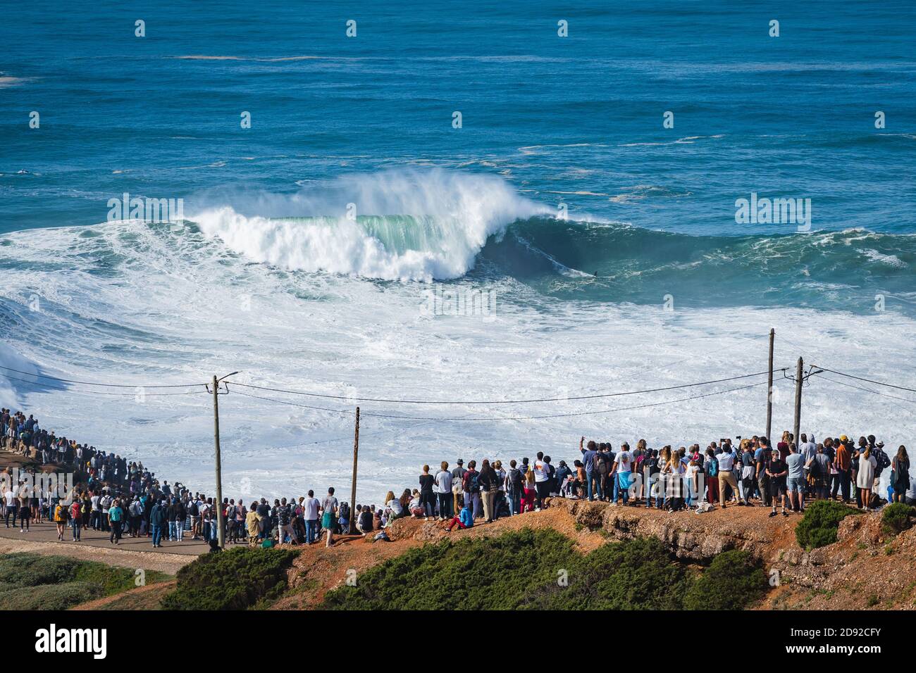 Menschen, die Big Wave Surfer beim Reiten der Riesenwelle am Strand Praia do Norte in Nazare, Portugal, beobachten. Stockfoto