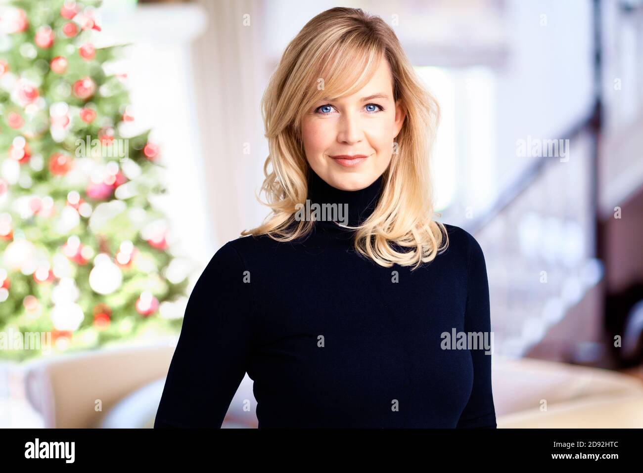 Porträtaufnahme einer lächelnden blonden Frau, die in ihrem Stehzimmer mit einem Weihnachtsbaum im Hintergrund steht. Stockfoto
