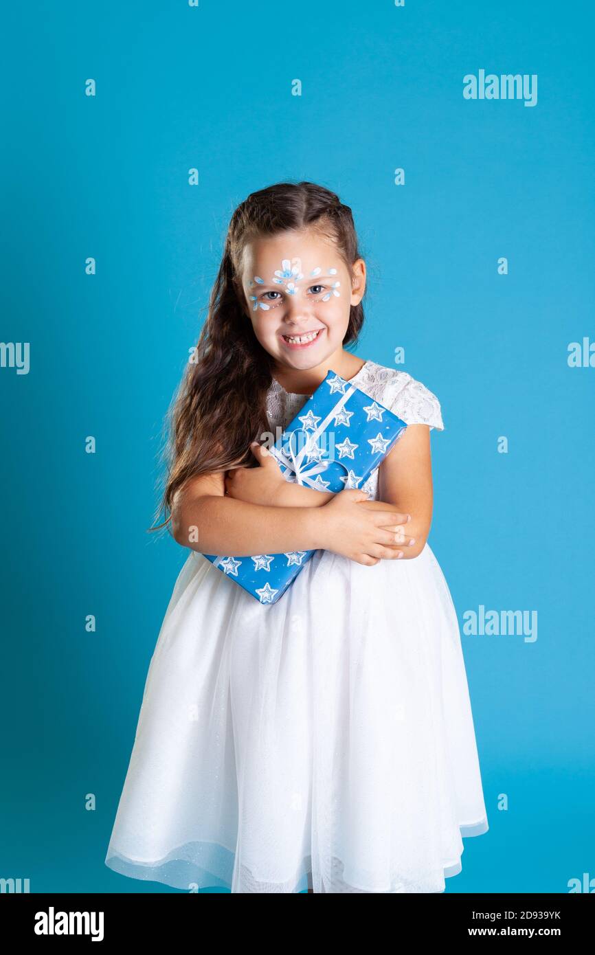 Eine kleine Prinzessin in einem weißen Kleid hält ein Weihnachtsgeschenk an ihre Brust, isoliert auf einem blauen Hintergrund Stockfoto