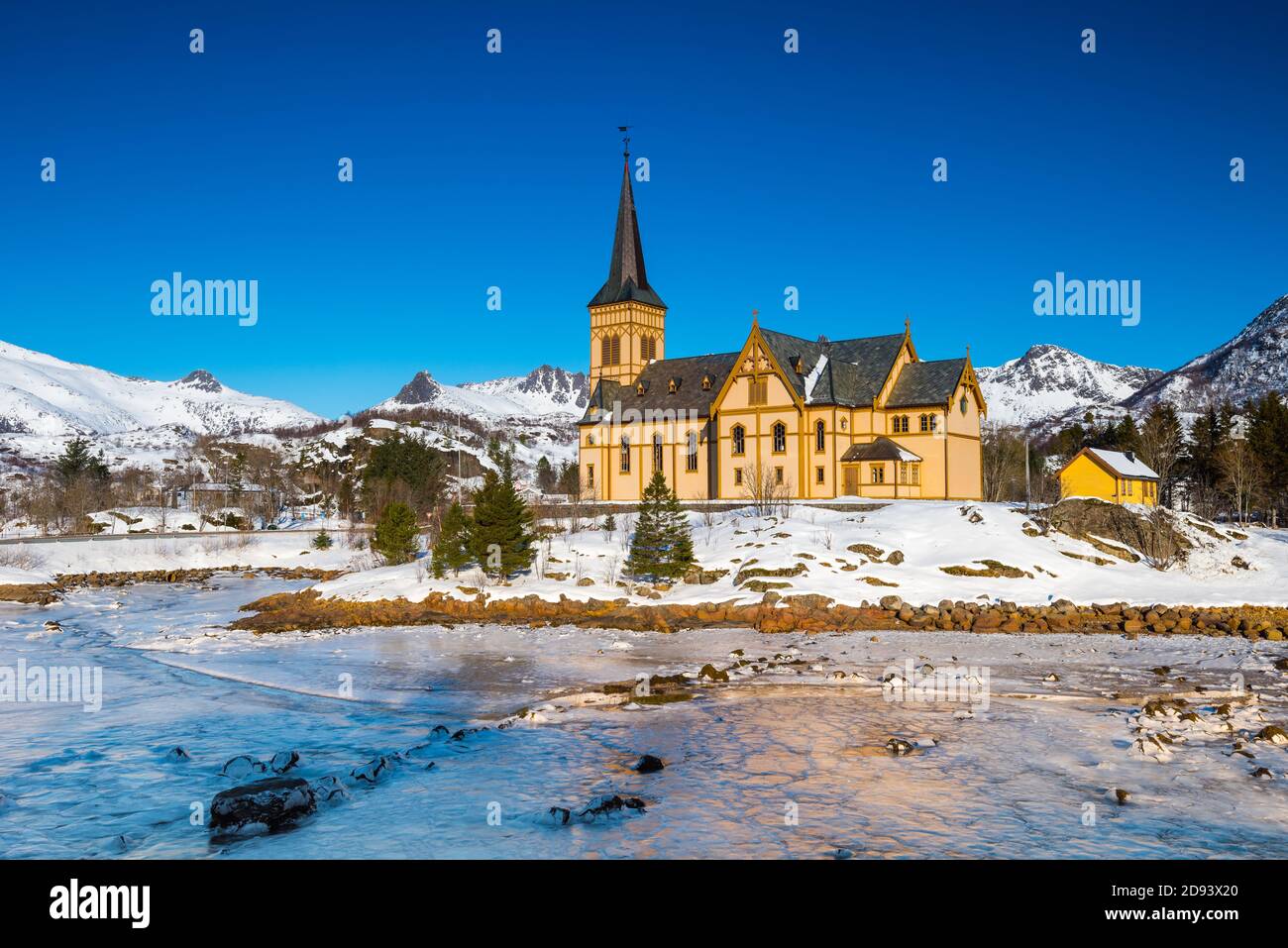 Die hölzerne gelbe Vågan Kirche am Strand auf dem Lofoten Inseln in Norwegen im Winter mit gefrorenen Fluss und Berge im Morgenlicht Stockfoto