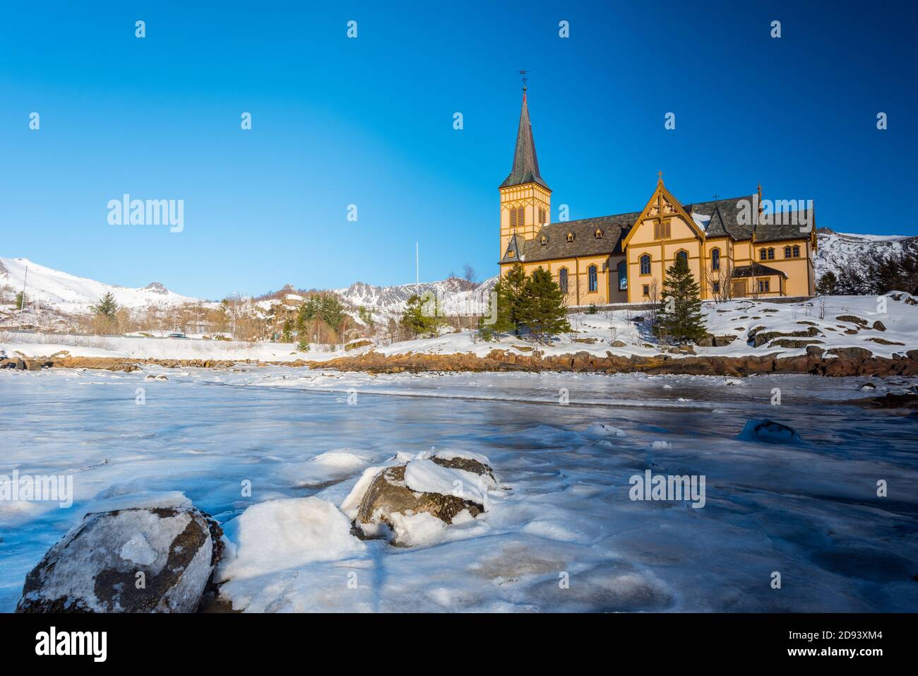 Die hölzerne gelbe Vågan Kirche am Strand auf dem Lofoten Inseln in Norwegen im Winter mit gefrorenen Fluss und Berge im Morgenlicht Stockfoto