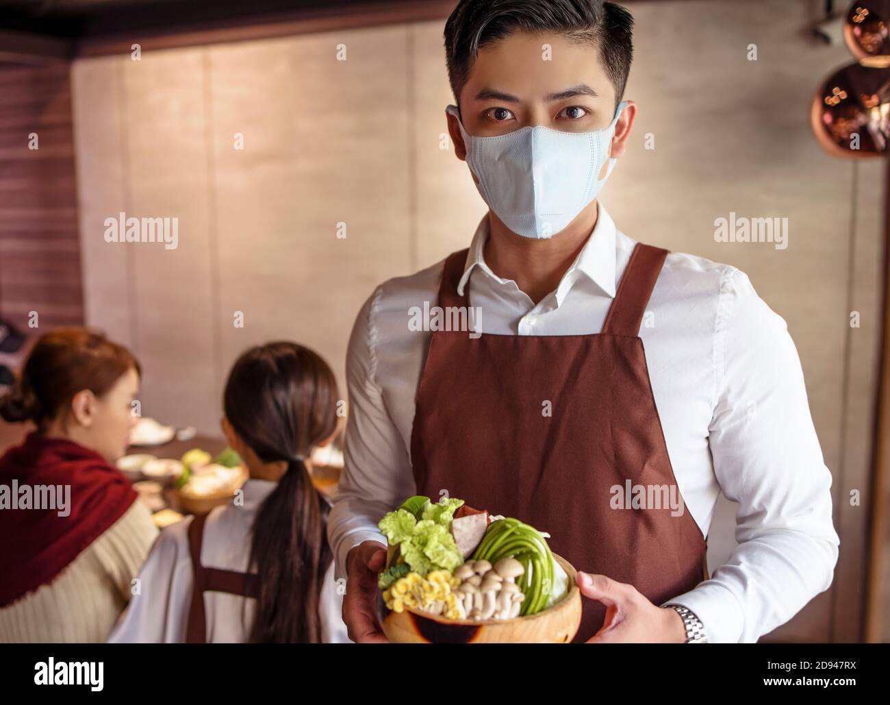 Junger Kellner trägt eine schützende Gesichtsmaske, während er Essen serviert Kunde im Restaurant Stockfoto