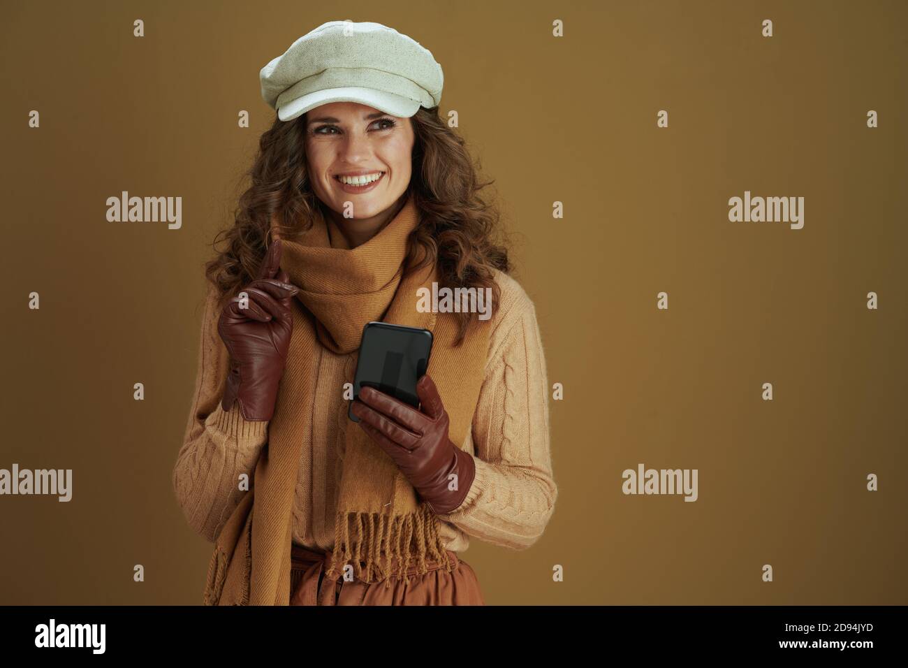Hallo Herbst. Glückliche moderne Frau im Schal mit Lederhandschuhen mit Smartphone-App auf Bronze Hintergrund. Stockfoto