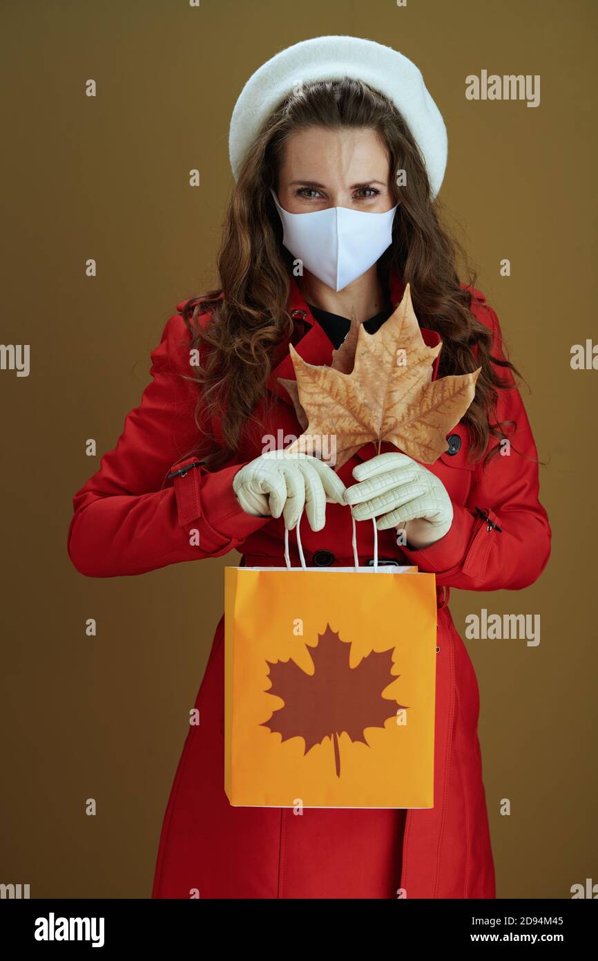 Leben während covid-19 Pandemie. Trendige Frau in rotem Mantel mit gelben Herbst Ahornblatt und Papier Einkaufstasche isoliert auf beigem Hintergrund. Stockfoto