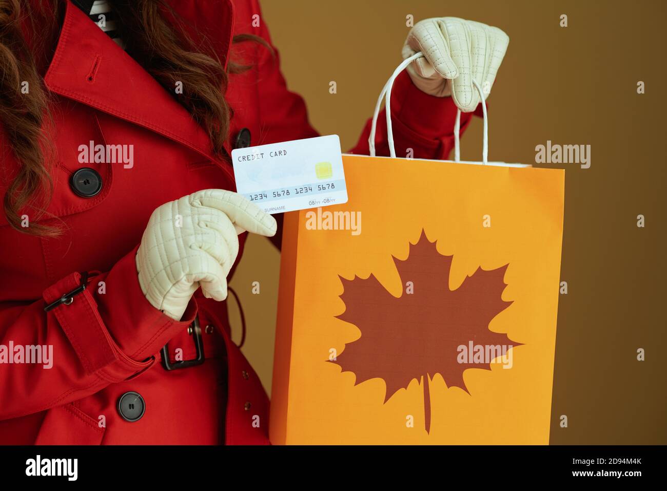 Hallo Herbst. Nahaufnahme einer Frau im roten Mantel mit Kreditkarte und Papier-Einkaufstasche vor bronzefarbenem Hintergrund. Stockfoto