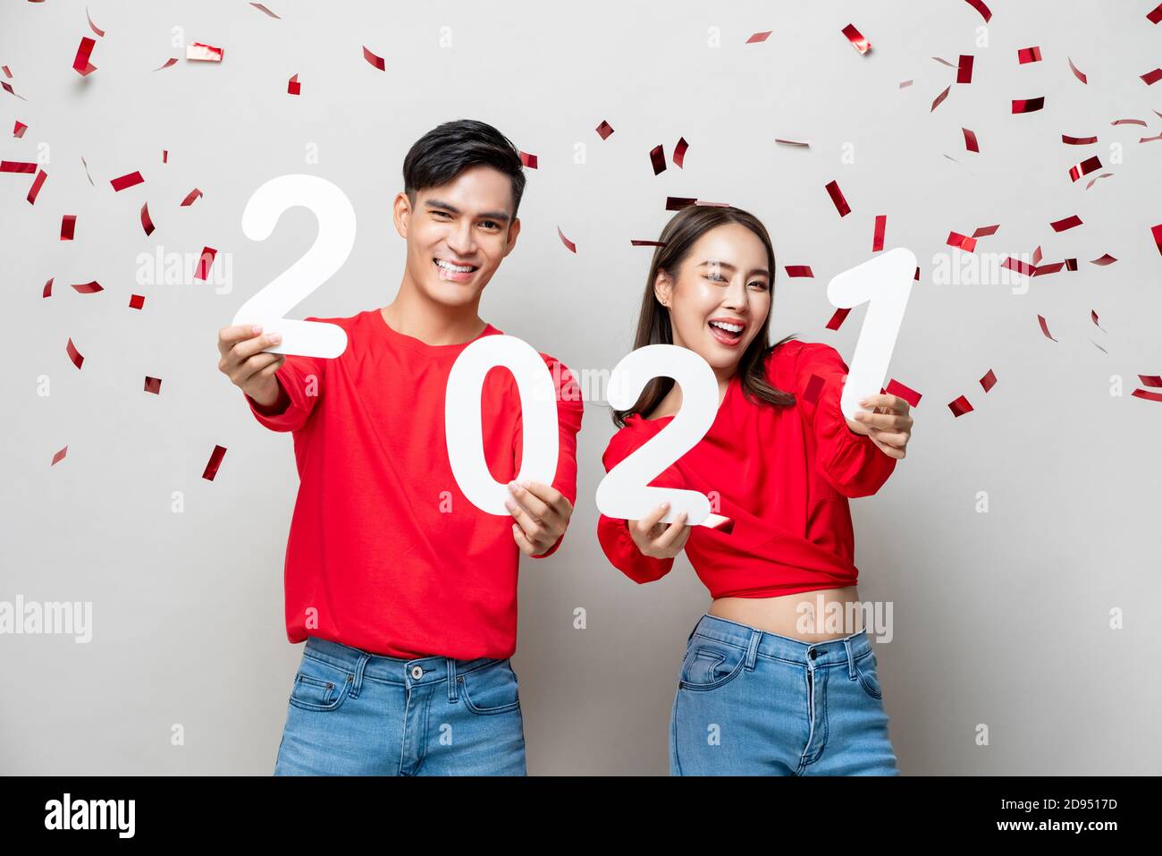 Lächelndes glückliches asiatisches Paar in roter legerer Kleidung mit 2021 Zahlen für das Jahreskonzept auf hellgrauem Hintergrund mit Konfetti Stockfoto