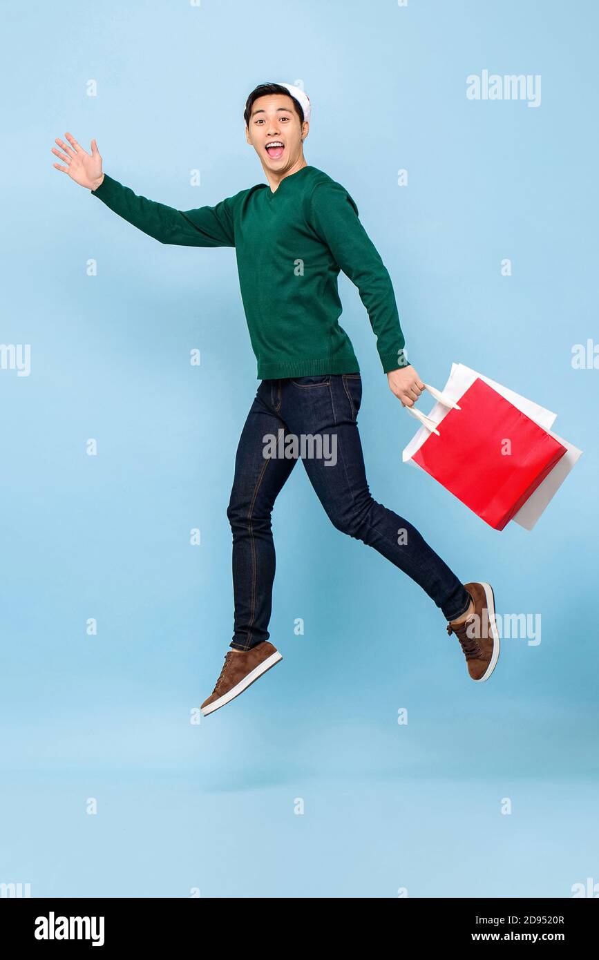 In voller Länge Porträt von glücklich junge gut aussehende asiatische Mann trägt weihnachtsmütze springen, während Einkaufstaschen isoliert auf Licht halten Blaue Studio-Backgr Stockfoto
