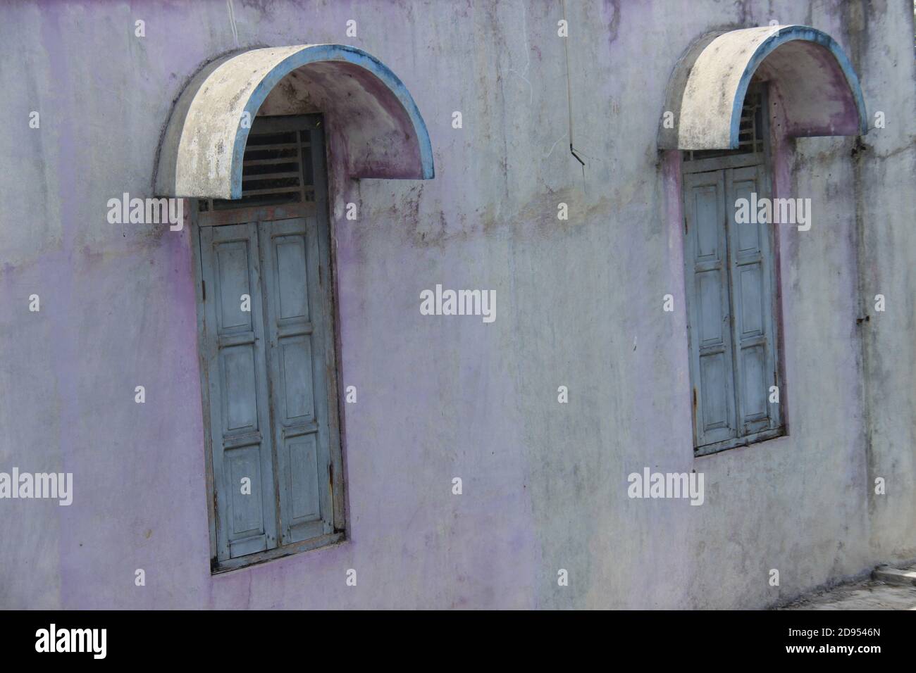 Altes Haus mit zwei blauen Holzfenstern und einer kaputten Wand. Altes Haus im ländlichen indonesien. Moosige Wandstruktur für den Hintergrund Stockfoto