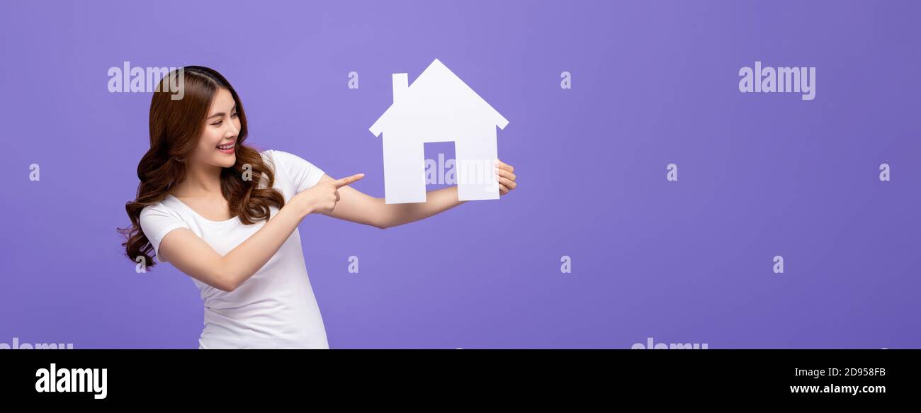 Glücklich schöne asiatische Frau lächelnd und zeigt auf Haus Ausschnitt Modell isoliert auf violettem Studio Banner Hintergrund mit Kopierraum Stockfoto