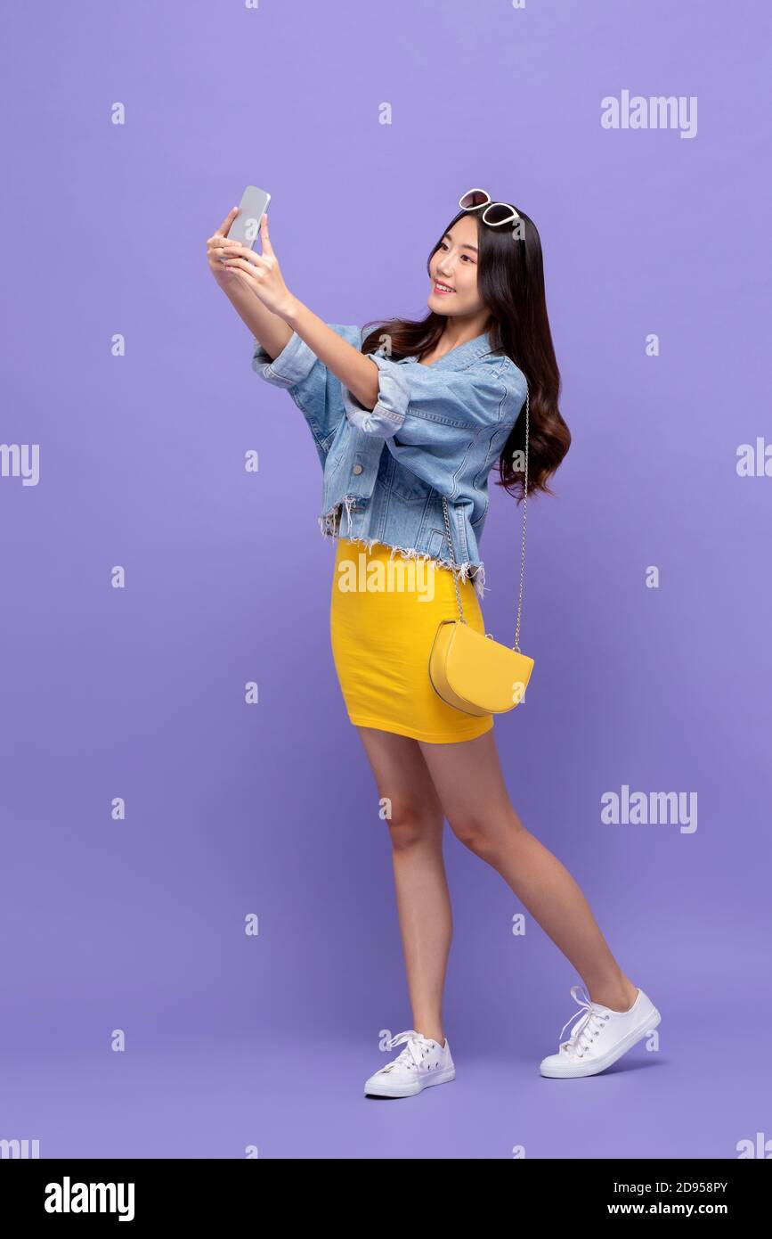 In voller Länge Porträt von lächelnd junge hübsche asiatische Frau nehmen Selfie mit Smartphone in isolierten Studio lila Hintergrund Stockfoto