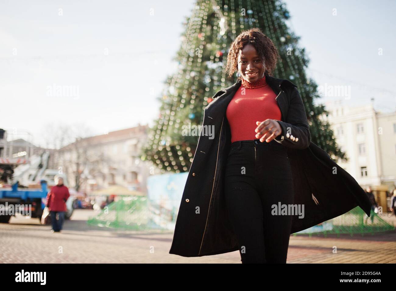 Porträt einer lockigen afrikanisch Frau tragen modischen schwarzen Mantel und roten Rollkragen posiert im Freien gegen Haupt-Neujahr Baum der Stadt. Stockfoto