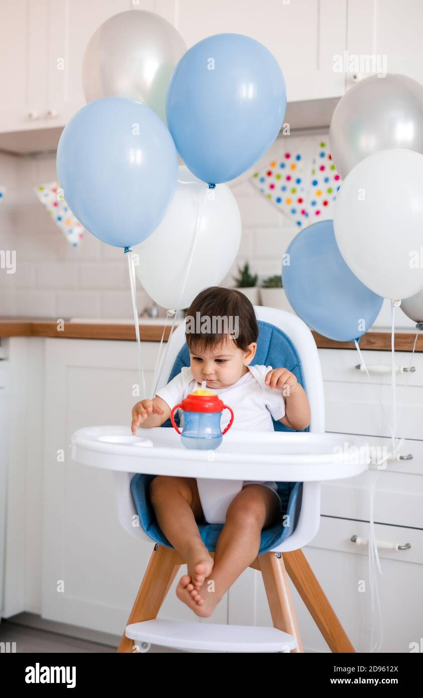 Kleiner Junge sitzt im Hochstuhl zu Hause auf Weiße Küche und Trinkwasser aus sippy Tasse auf Hintergrund Mit Luftballons Stockfoto