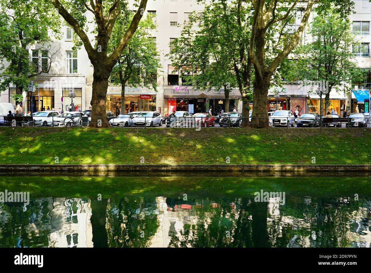 Die berühmte Einkaufsmeile Königsallee: Blick über den grünen Stadtkanal 'Kö-Graben' von der 'ruhigen' Seite auf die 'belebte Seite' mit vielen Geschäften. Stockfoto