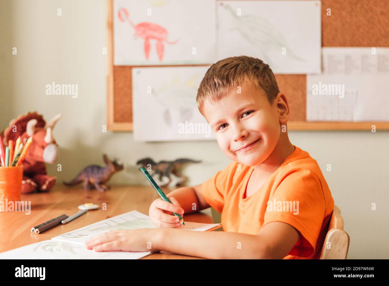 Nettes Kind Junge Zeichnung Dinosaurier auf Bild mit Buntstiften Sitzen am Schreibtisch in seinem Zimmer Stockfoto