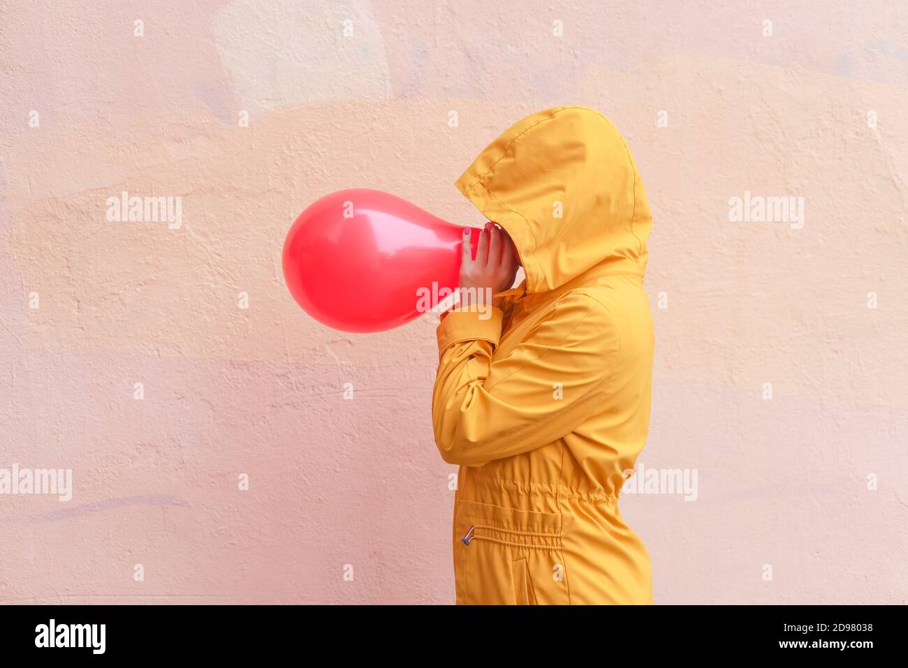 Person in gelbem Mantel mit Kapuze mit rotem Ballon an beigefarbener Wand. Abstraktes konzeptuelles Porträt Stockfoto