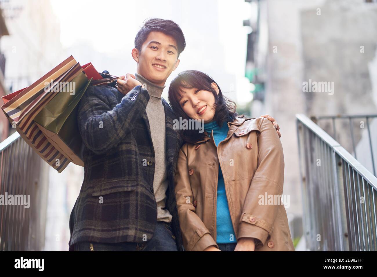 Außenportrait eines glücklichen jungen asiatischen Pärchen, das von einem Einkaufsbummel zurückkehrt und lächelnd auf die Kamera schaut Stockfoto