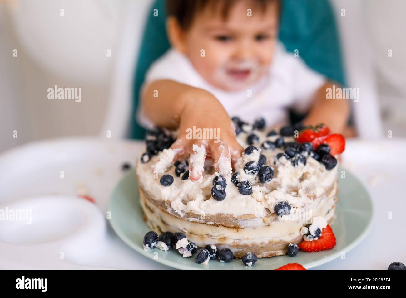 Kleiner Junge, der in einem Hochstuhl in der weißen Küche sitzt Und Verkostung ersten Jahr Kuchen mit Früchten auf Hintergrund mit Ballons Stockfoto