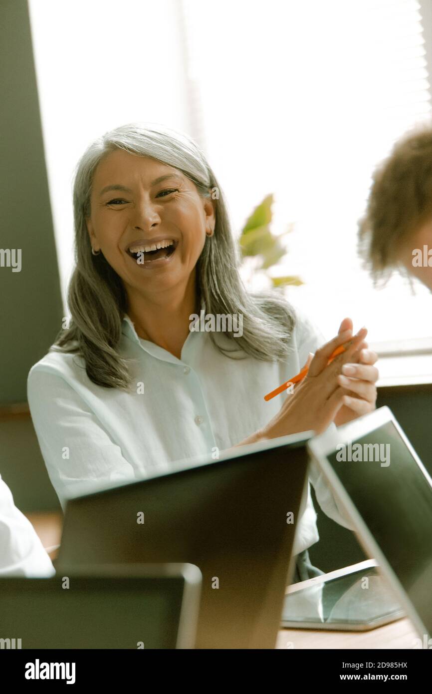 Lustige asiatische ältere Frau lacht bei Geschäftstreffen sitzen an Schreibtisch mit Laptop-Computern am Fenster Im Hintergrund Stockfoto