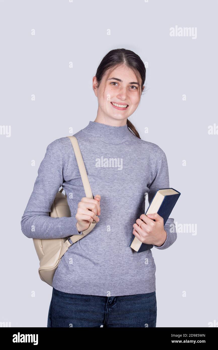 Selbstbewusstes Studentenmädchen lächelnd hält ein Buch und trägt ihren Rucksack. Fröhliche Student Mädchen mit positiven Gesichtsausdruck isoliert auf weißen BA Stockfoto