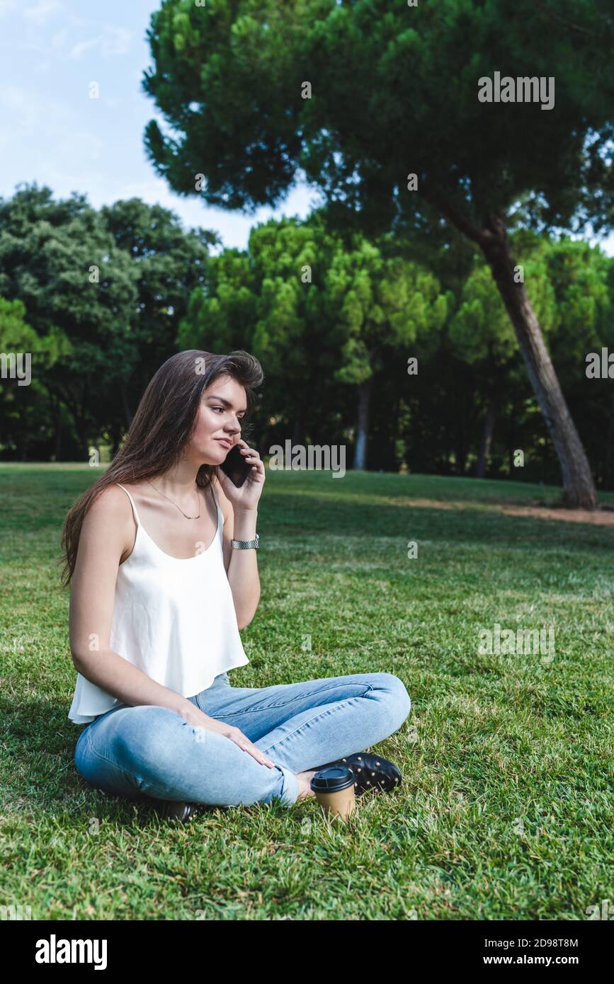 Breites vertikales Porträt einer jungen kaukasischen Frau mit casual Kleidung sitzt auf dem Gras eines grünen Parks mit Gekreuzte Beine und Telefonieren Stockfoto