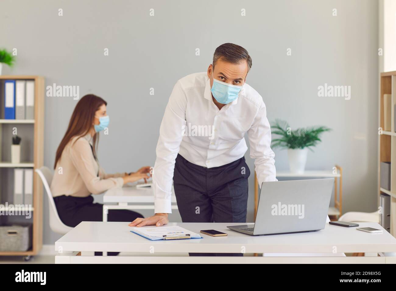 Erwachsener Mann in einer medizinischen Schutzmaske steht im Büro vor dem Hintergrund seines Kollegen. Stockfoto