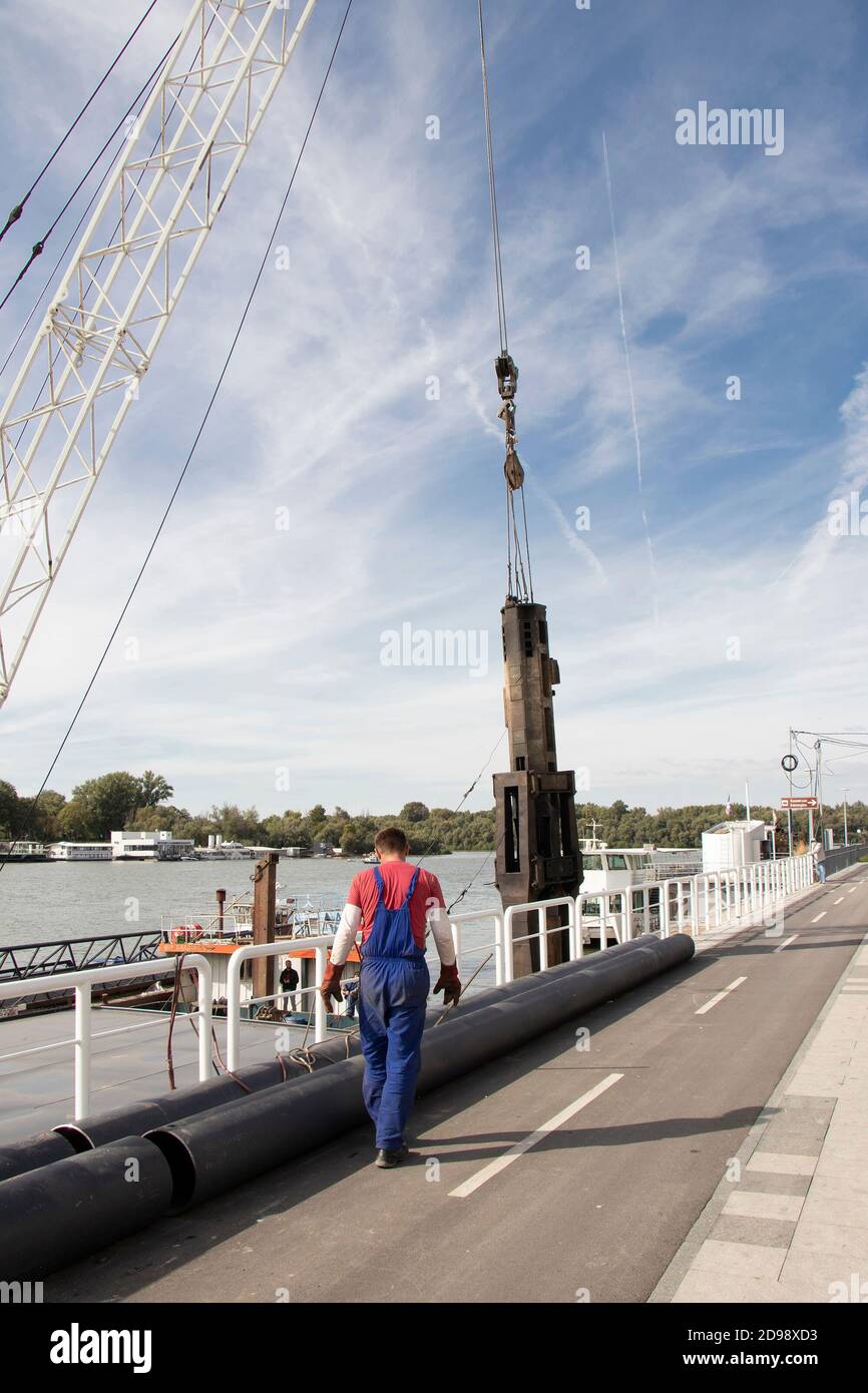Belgrad, Serbien - 9. Oktober 2020: Baukran, der einen großen industriellen Stahlspieß für den Flussponton und einen Arbeiter hebt, mit Blick auf das Flussufer Stockfoto