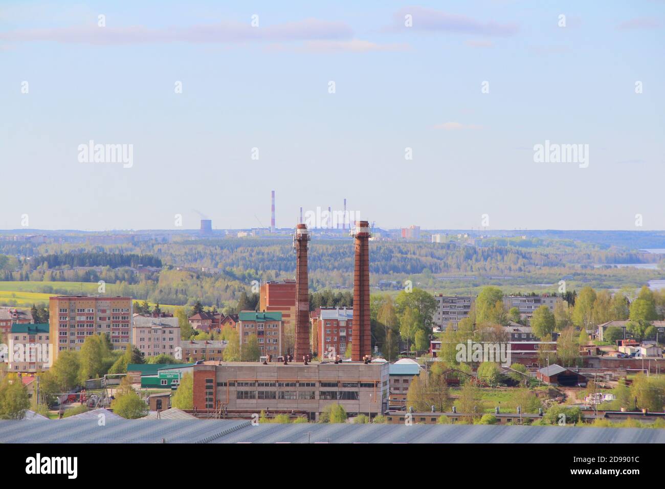 Die städtische Industrielandschaft mit Fabrik, Wohngebäuden und Rohren am Horizont im Sommer in Russland. Eine menschengemachte Landschaft. Eine Industriestadt mit grünen Bäumen. Das Konzept von Industrie, Ökologie und Umweltverschmutzung. Zwei Werksrohre im Vordergrund. Stockfoto