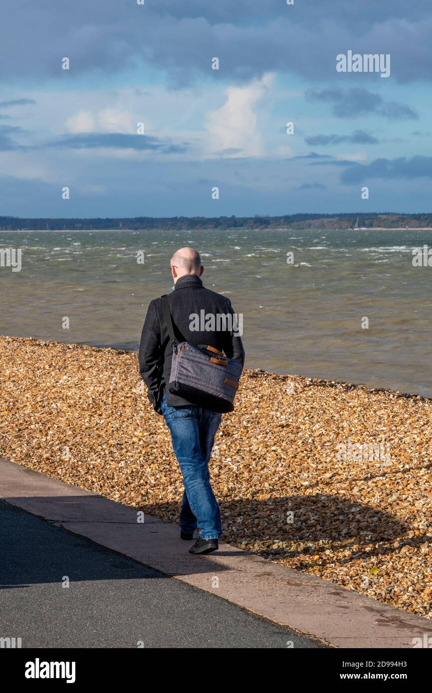 Ein Mann mit kahlem Kopf und einer Manntasche, der an einem im Herbst bewölkten Tag an einem Strand am Meer entlang läuft. Stockfoto