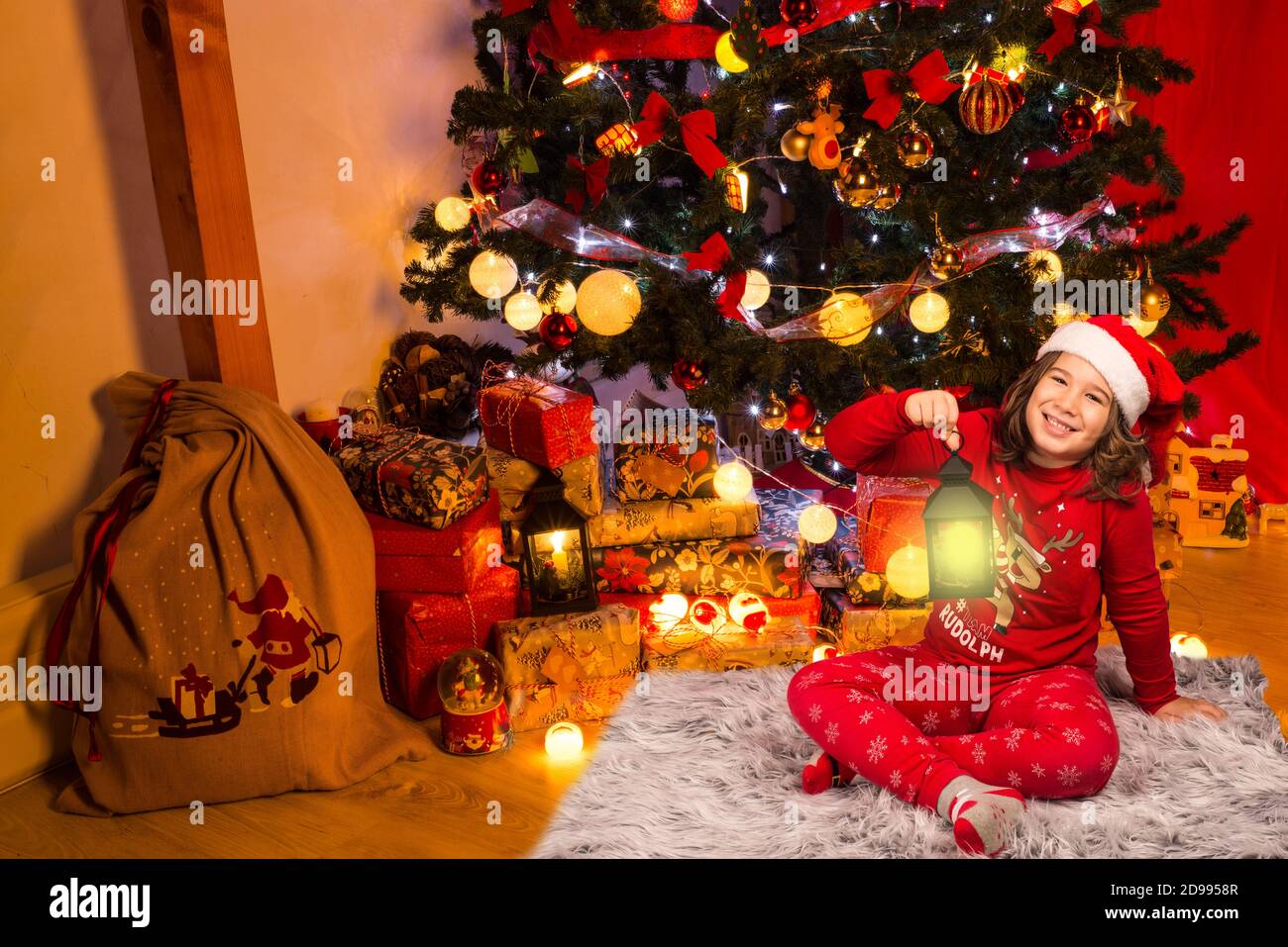 Glückliches Kind, das vor dem Weihnachtsbaum einen beleuchteten Laternenpfosten zeigt Mit Geschenken Stockfoto