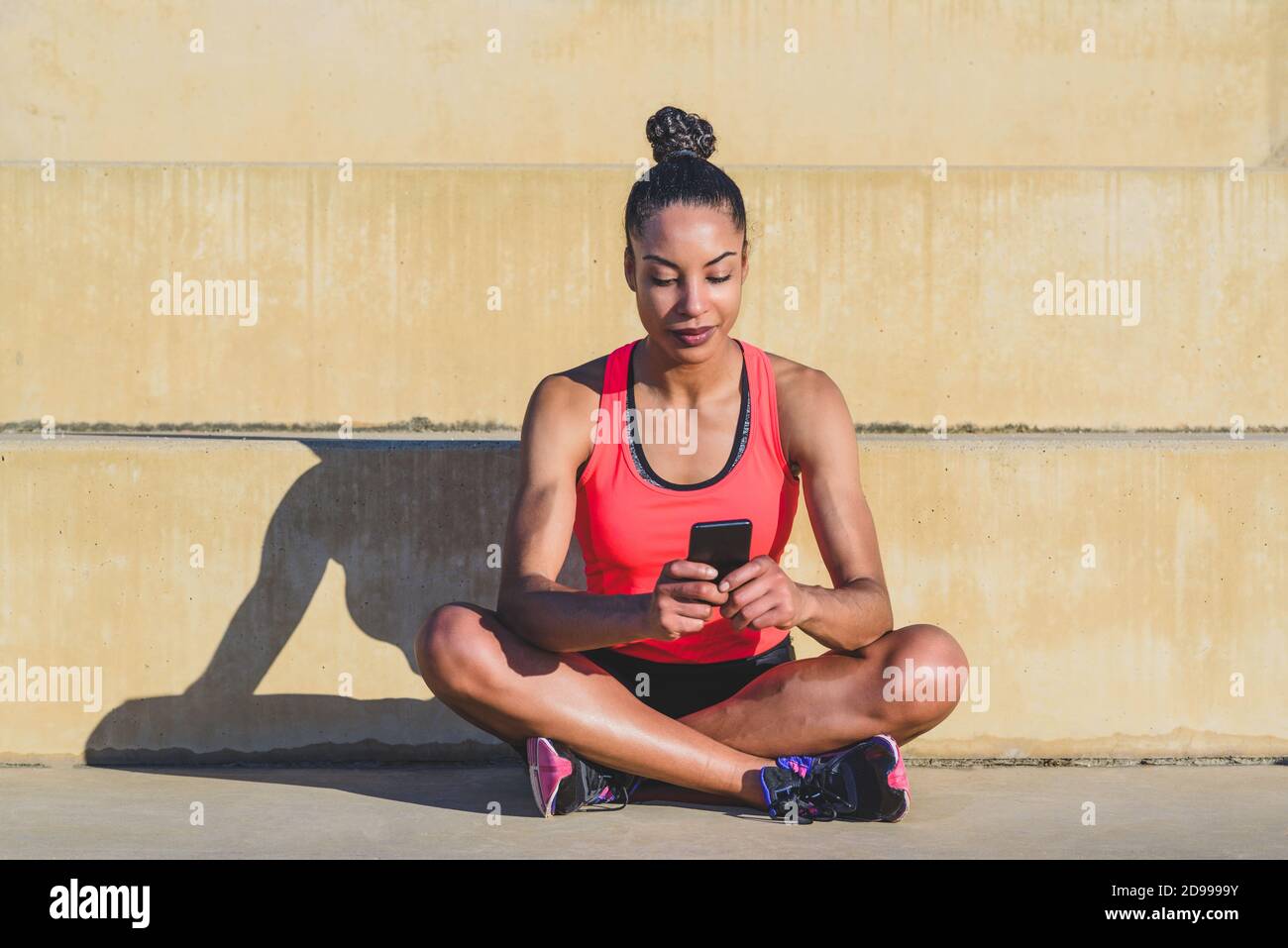 Attraktive junge afroamerikanische Frau mit ihren Haaren gebunden und Tragen Sportbekleidung sitzen gekreuzt auf Betonstufen mit ihrem Handy Telefon eingeschaltet Stockfoto