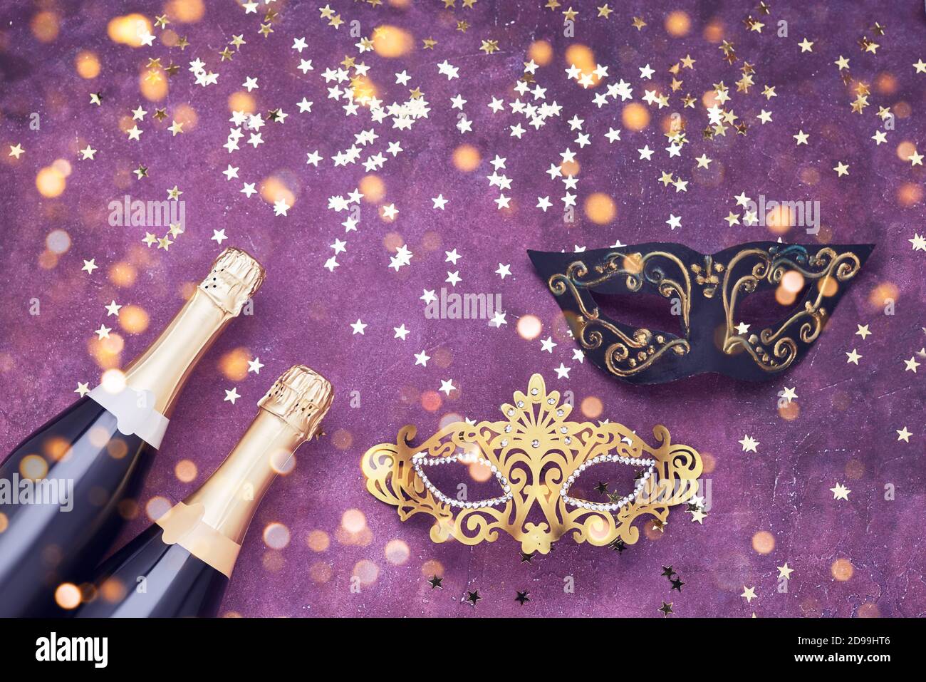 Zwei Champagnerflaschen, goldene und schwarze Karnevalsmaske und Konfetti-Sterne auf violettem Hintergrund. Flaches Lay von Weihnachten, Neujahr, Purim, Karneval Promi Stockfoto