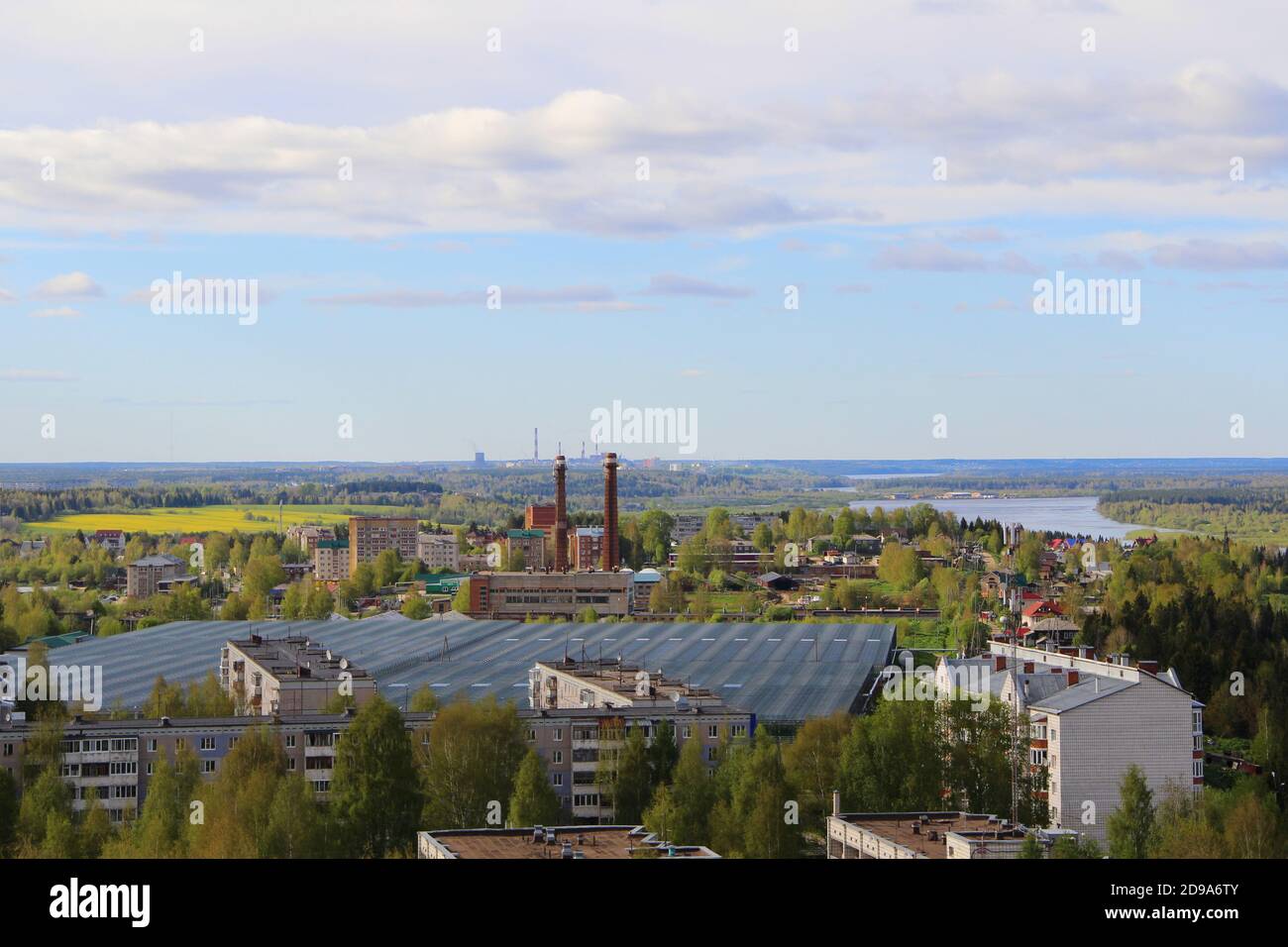 Die städtische Industrielandschaft mit Fabrik, Wohngebäuden und Rohren am Horizont im Sommer in Russland. Eine menschengemachte Landschaft. Eine Industriestadt mit grünen Bäumen. Das Konzept von Industrie, Ökologie und Umweltverschmutzung. Zwei Werksrohre im Vordergrund. Stockfoto